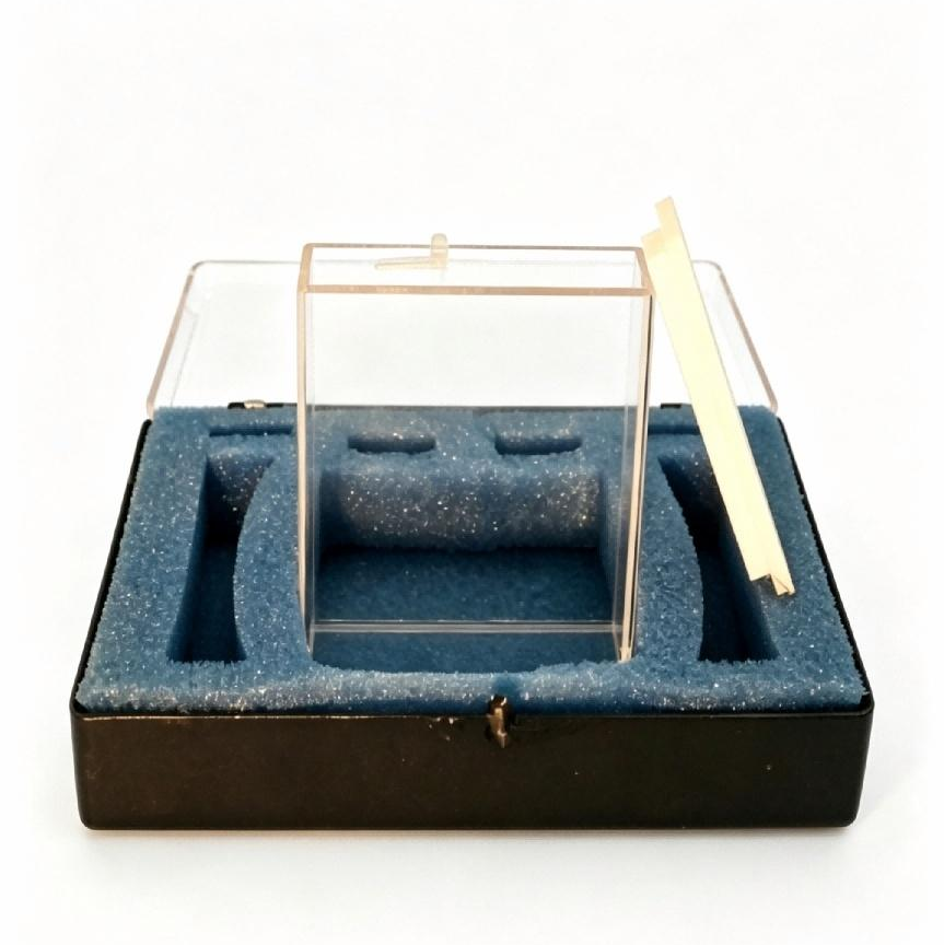 A clear acrylic jewelry display stand with a hinged lid, set inside a black presentation box with blue foam insert and a small cream-colored tag, photographed on a white background.
