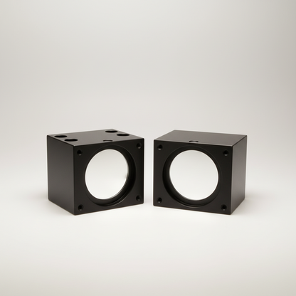 Two black square metal components with circular silver reflective surfaces, placed on a light wood surface.