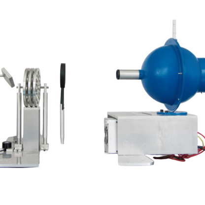 A complex scientific apparatus or miniature machine setup on a reflective green surface against a plain white wall. The setup includes a blue spherical component on a metal base, a silver cylindrical component with a black handle, and a circular metal wheel assembly on a stand.